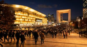 Façade illuminée de la Plénitude Arena à Nanterre lors d'un concert de Céline Dion, avec une foule de spectateurs arrivant depuis l'esplanade de La Défense et la Grande Arche visible en arrière-plan