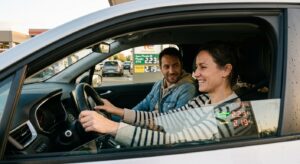 Deux covoitureurs souriants dans une voiture sur une aire d'autoroute française, panneau de prix carburant visible à 2,20 €/L en arrière-plan, illustrant la hausse du recours au covoiturage en 2026